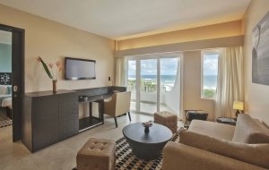 der wohnbereich einer grossen schönen suite mit couch fernsehtisch und einem smart tv fernseher an der wand die offene tür gewährt einen blick in das schlafzimmer und die grosse terrassentür an der aussenwand einen blick auf den balkon und den strand am m